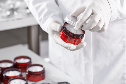 Factory worker in white gown and gloves holding red container with skin care product touching it with finger testing quality. Representing new product on factory. Showing high quality beauty mask